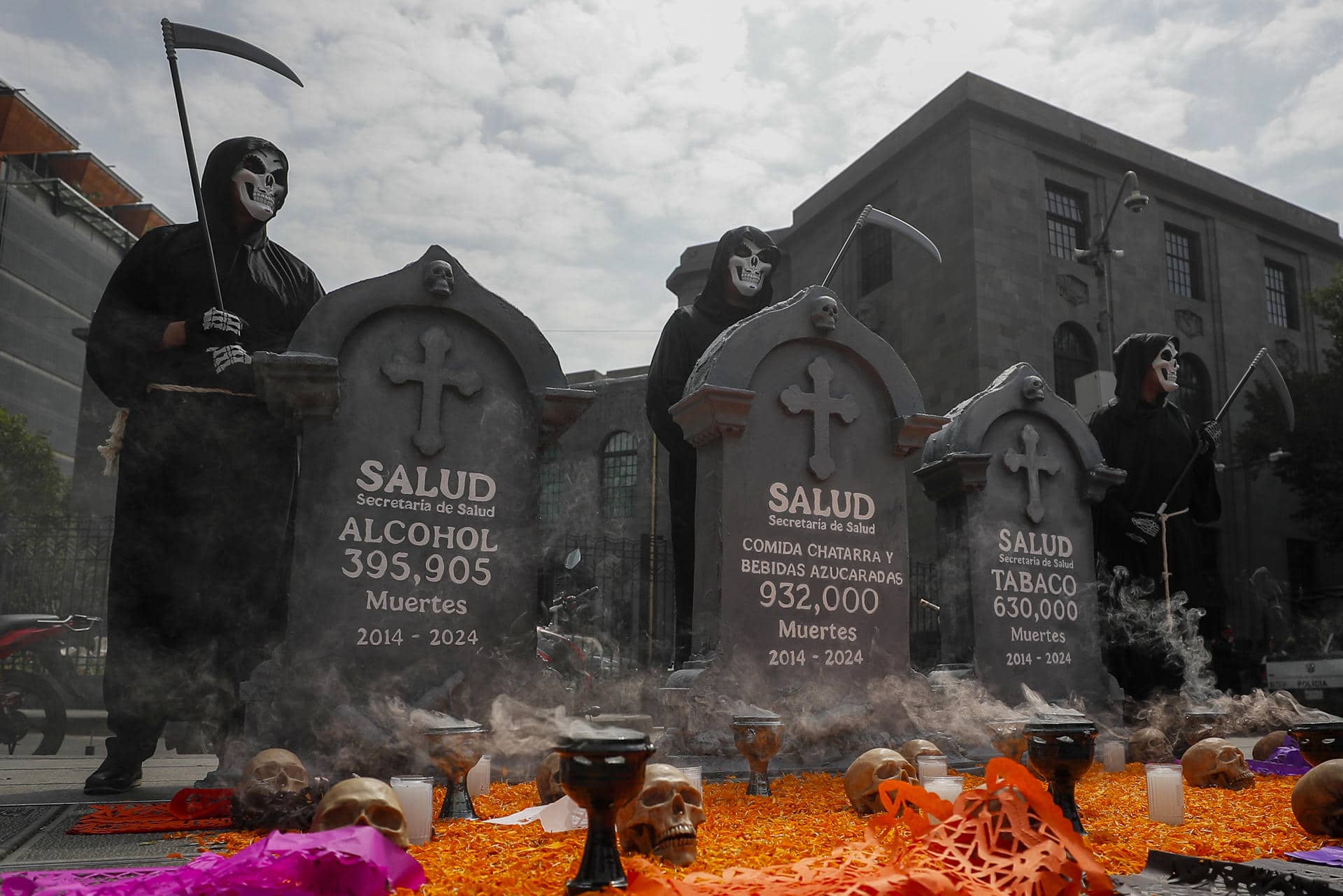 Miembros de organizaciones sociales disfrazados de calaveras posan junto a una ofrenda con esquelas que muestran presuntas cifras de muertes por alcohol, comida chatarra y bebidas azucaradas y tabaco en los últimos 10 años, que colocaron frente a la Secretaría de Salud este jueves, en la Ciudad de México (México). EFE/ Isaac Esquivel