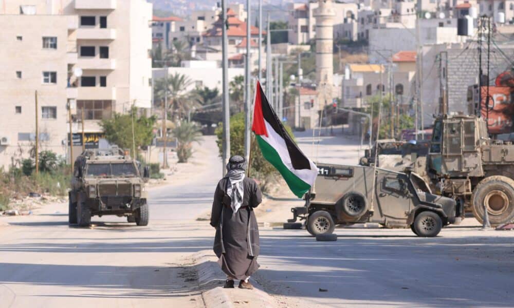 Un hombre lleva una bandera palestina mientras vehículos militares israelíes avanzan por una calle durante una redada llevada a cabo por fuerzas israelíes en la ciudad cisjordana de Tulkarem, este jueves. Según el Ministerio de Salud palestino en Cisjordania, al menos un palestino murió y otros cinco resultaron heridos durante la redada en Tulkarem y su campo de refugiados.EFE/ Alaa Badarneh