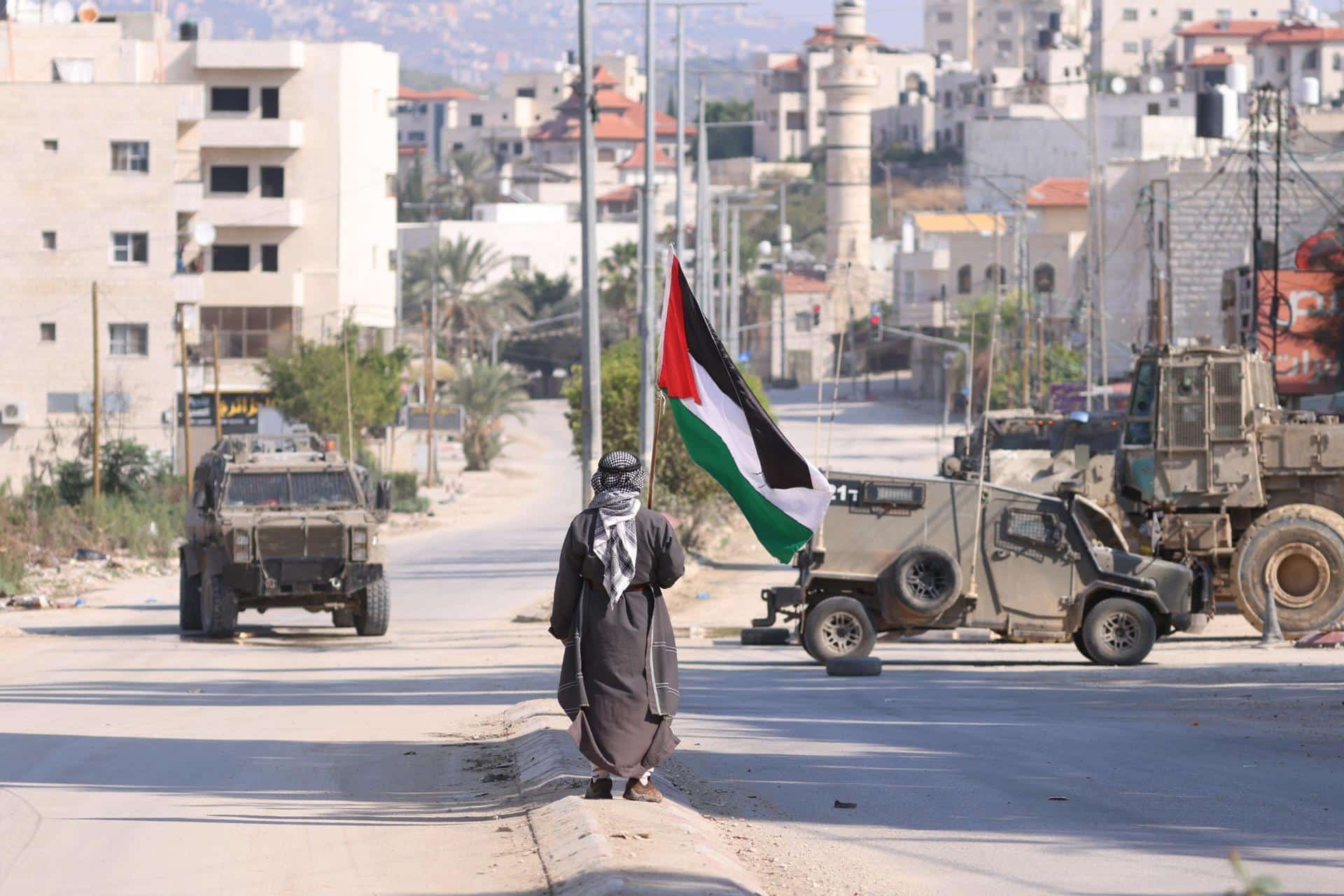 Un hombre lleva una bandera palestina mientras vehículos militares israelíes avanzan por una calle durante una redada llevada a cabo por fuerzas israelíes en la ciudad cisjordana de Tulkarem, este jueves. Según el Ministerio de Salud palestino en Cisjordania, al menos un palestino murió y otros cinco resultaron heridos durante la redada en Tulkarem y su campo de refugiados.EFE/ Alaa Badarneh