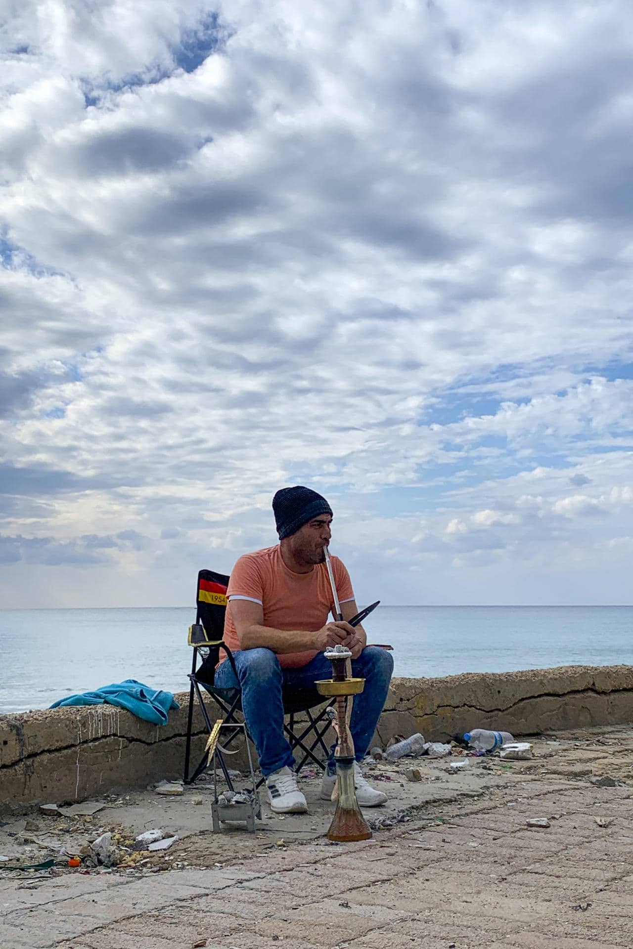 Un hombre desplazado fuma una pipa de agua sentado en su silla plegable en el paseo marítimo de Tiro, en el sur del Líbano, mientras observa a los equipos de la Defensa Civil que ya comienzan a retirar escombros de la ciudad, adonde ha regresado incapaz de volver aún a su aldea cerca de la divisoria con Israel. EFE/Noemí Jabois