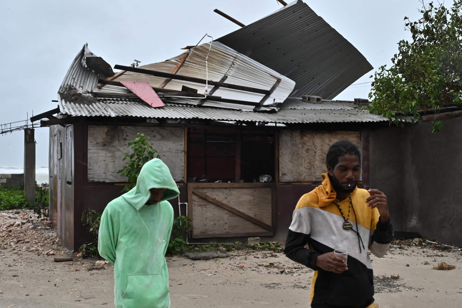 Personas pasan frente a una casa afectada por los vientos preliminares del huracán Melissa este lunes, en la playa pesquera de Hellshire, en Portmore (Jamaica). EFE/Rudolph Brown