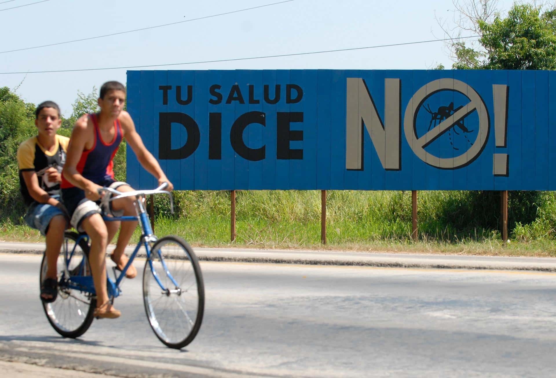 Fotografía de archivo de un par de jóvenes sobre una bicicleta al pasar frente a un cartel de campaña contra el Dengue, en Alamar (Cuba). EFE/Orlando Barría
