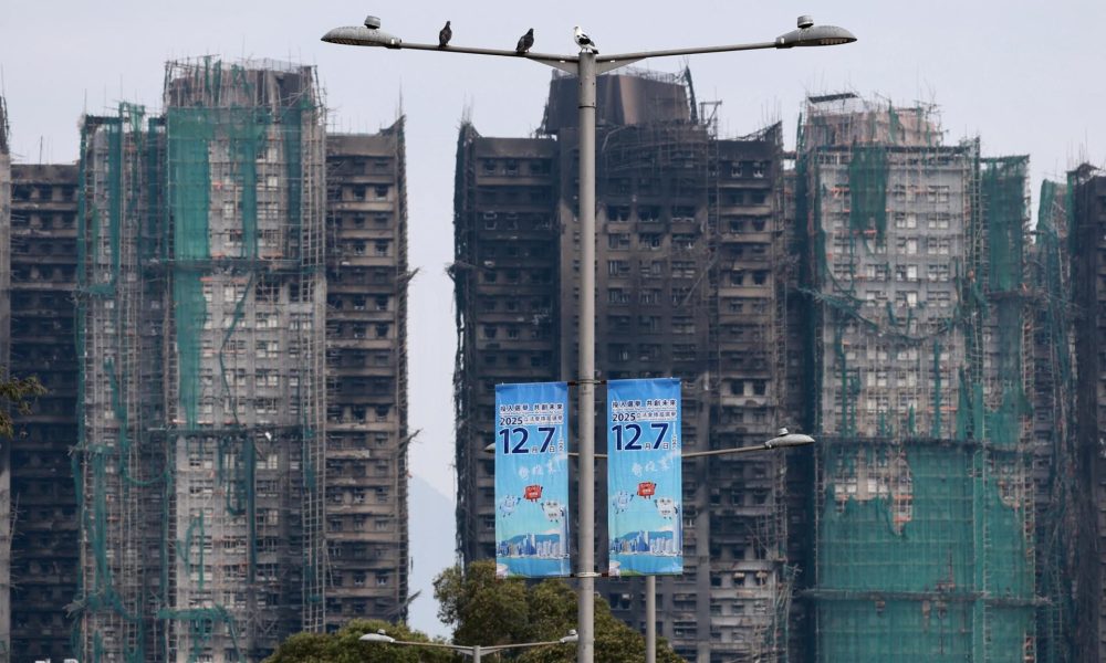HONG KONG (China), 07/12/2025.- Los restos calcinados del Wang Fuk Court se alzan tras pancartas promocionales cerca de un colegio electoral en el distrito de Tai Po durante las elecciones generales al Consejo Legislativo en Hong Kong, China, el 7 de diciembre de 2025. (Elecciones) EFE/EPA/MAY JAMES