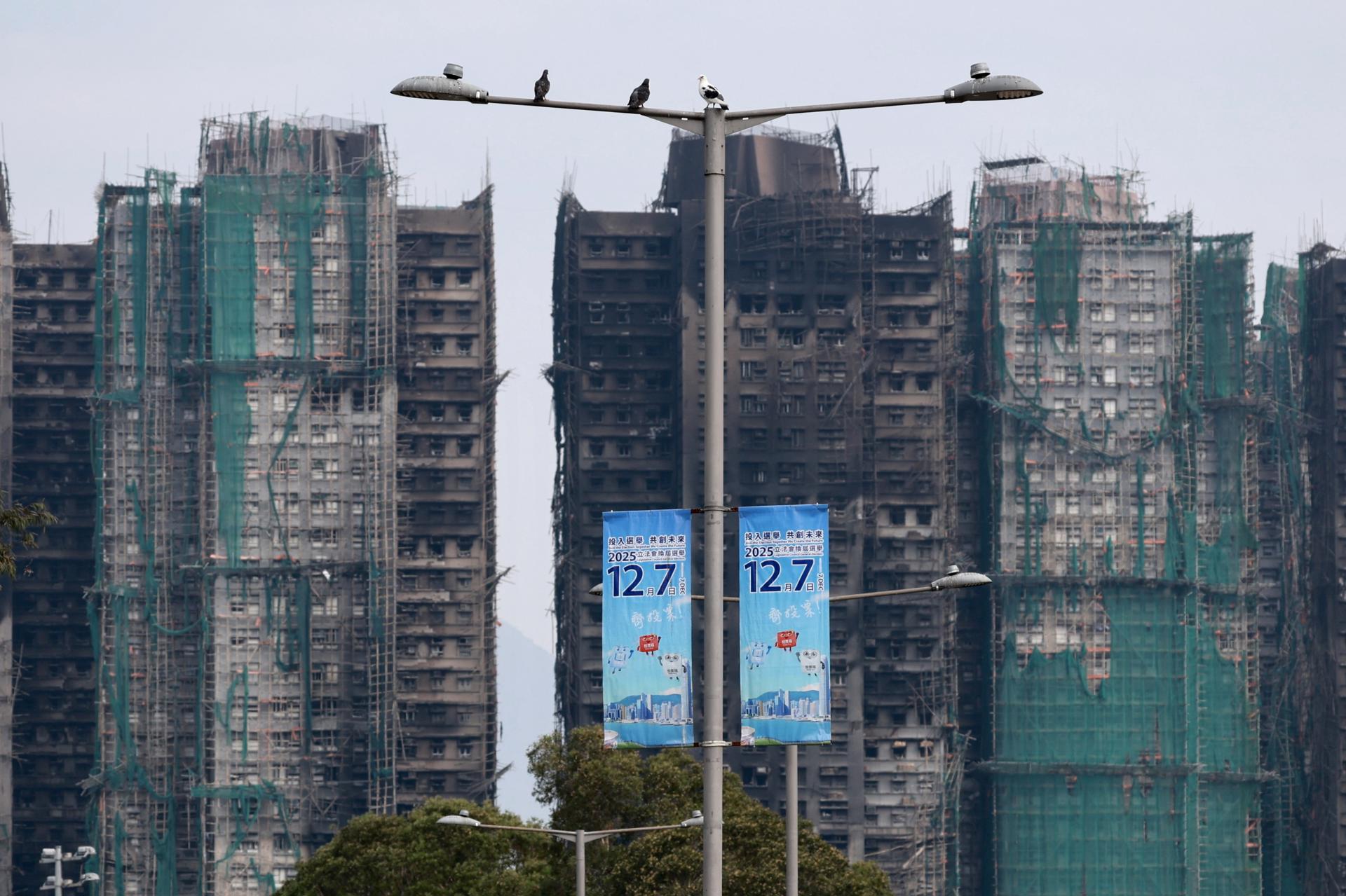HONG KONG (China), 07/12/2025.- Los restos calcinados del Wang Fuk Court se alzan tras pancartas promocionales cerca de un colegio electoral en el distrito de Tai Po durante las elecciones generales al Consejo Legislativo en Hong Kong, China, el 7 de diciembre de 2025. (Elecciones) EFE/EPA/MAY JAMES