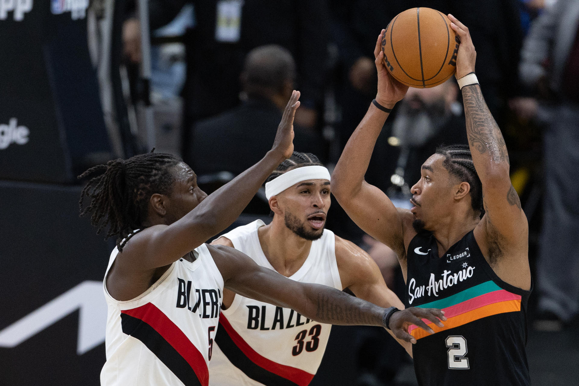 Dylan Harper (d) de Spurs recibe la marca de Jrue Holiday (i) y Toumani Camara de Trail Blazers durante el juego de este domingo en el AT&T Center de San Antonio (Texas. EFE/ Carlos Ramírez